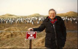 Harold Briley en una postal de las Falklands, junto a un cartel advirtiendo de un campo minado y en el fondo una colonia de pingüinos